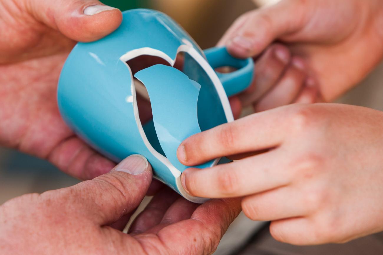 Zwei Hände halten eine zerbrochene, blaue Tasse mit herausgebrochenem Stück.