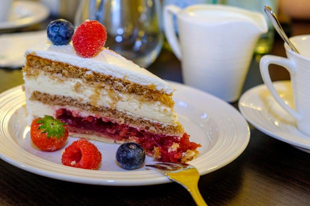 Nahaufnahme eines Stücks Beerentorte auf einem weißen Teller, garniert mit frischen Himbeeren, Blaubeeren und einer Erdbeere in einem Café-Setting mit Tassen und Milchkännchen im Hintergrund.