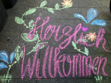Bunte Kreidezeichnung „Herzlich Willkommen“ mit Blumen auf Gehweg.