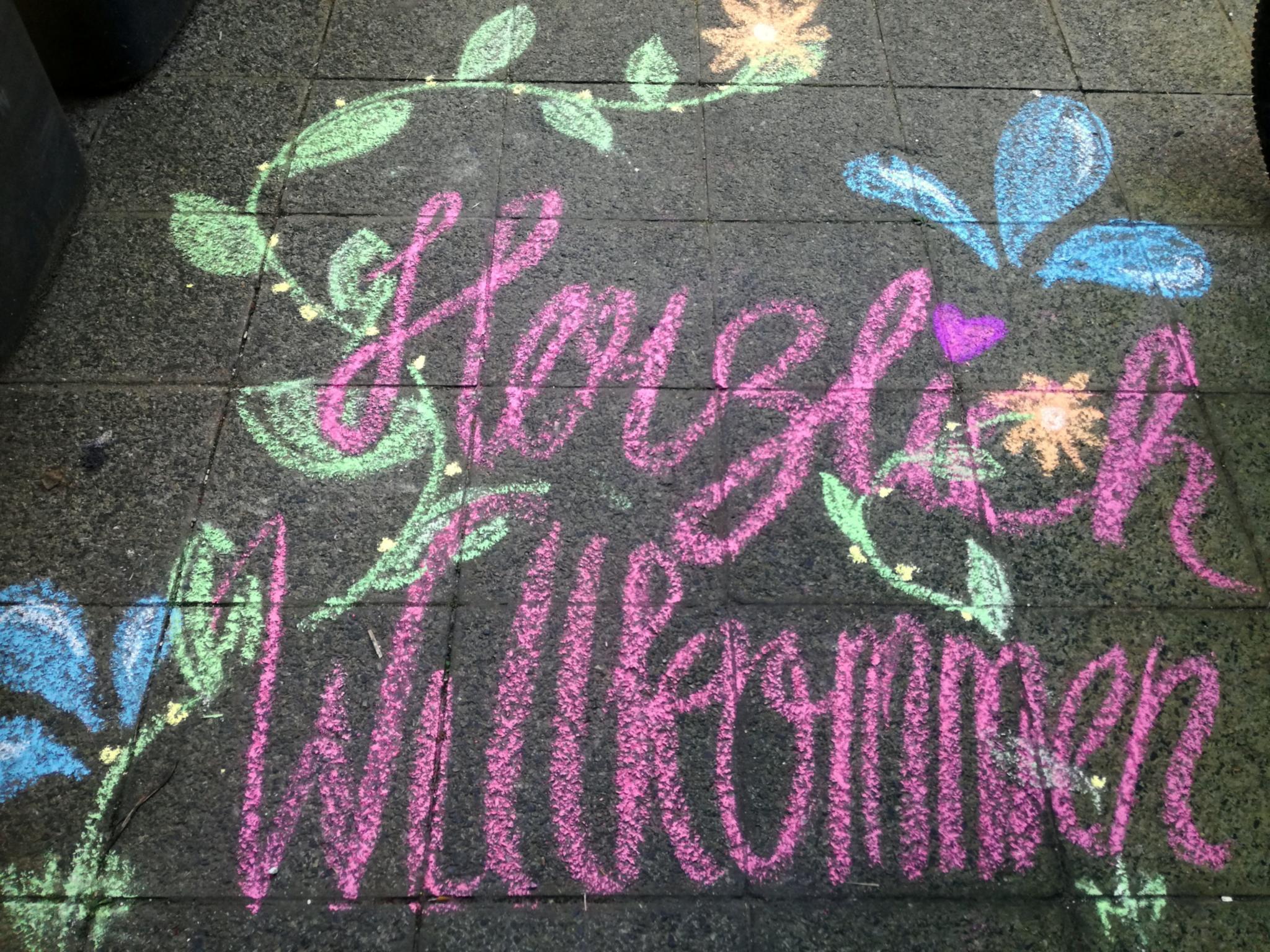 Bunte Kreidezeichnung „Herzlich Willkommen“ mit Blumen auf Gehweg.