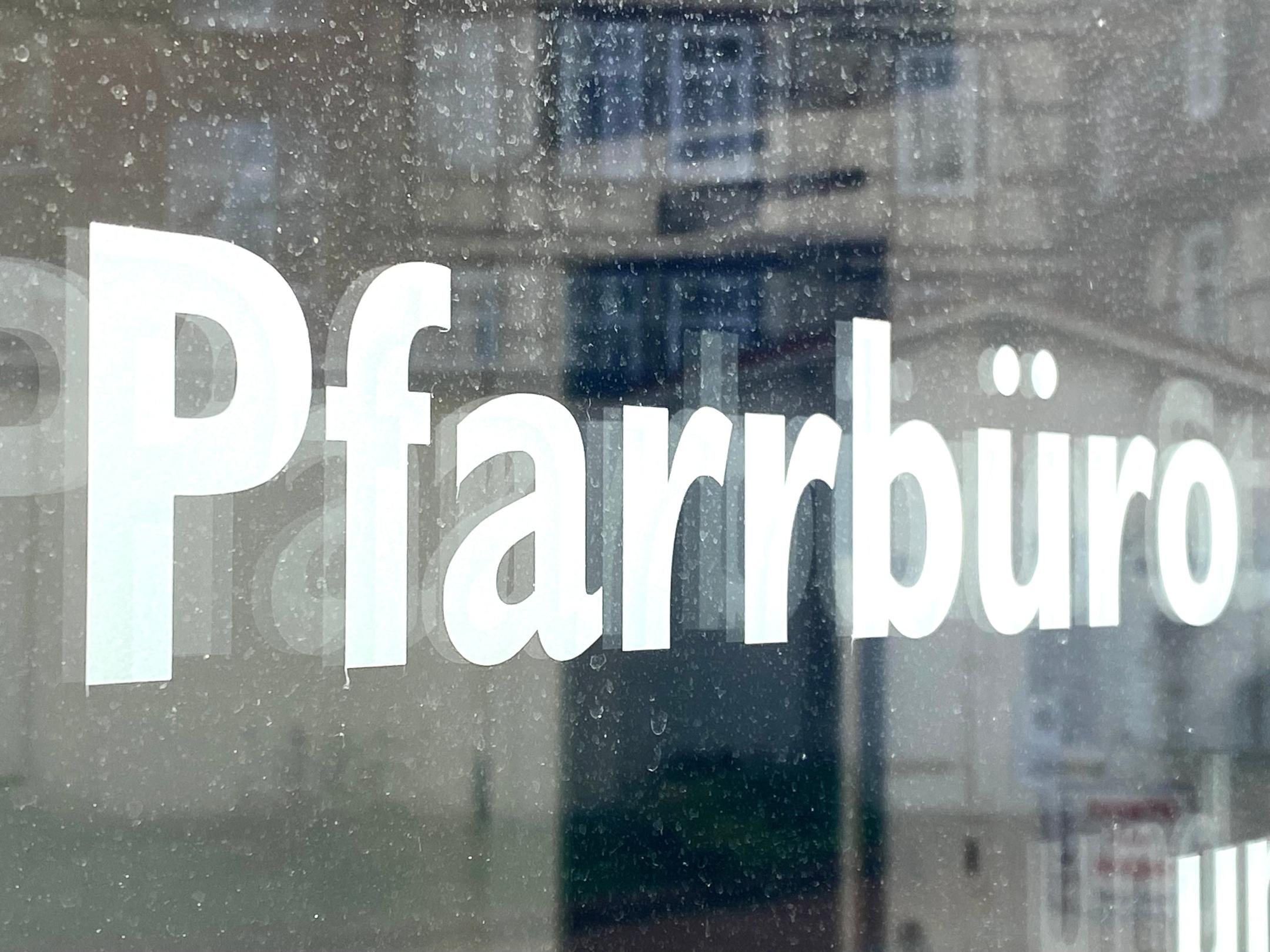 Weiße Schrift „Pfarrbüro“ auf einer Glasscheibe mit Spiegelungen.