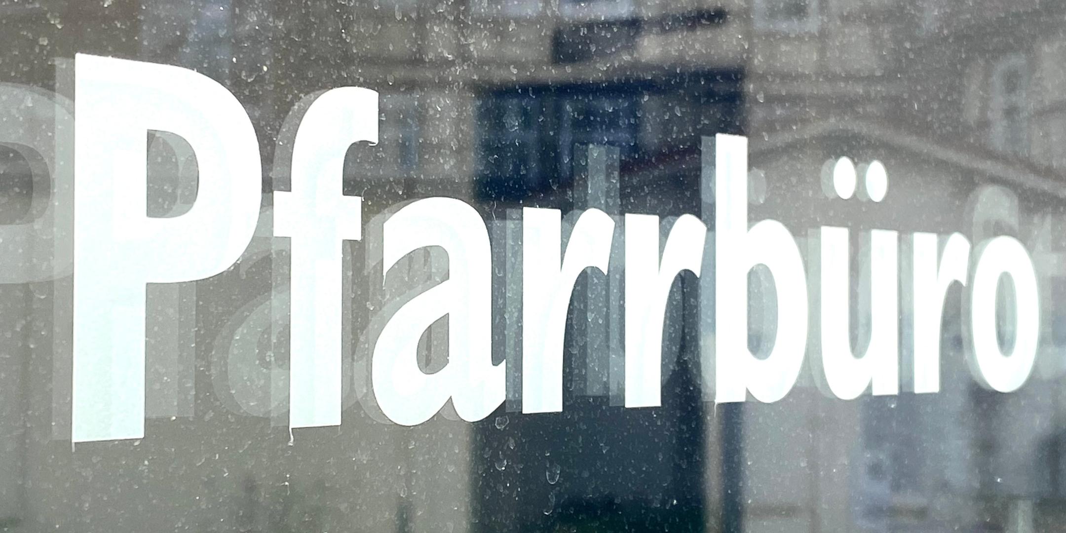 Weiße Schrift „Pfarrbüro“ auf einer Glasscheibe mit Spiegelungen.