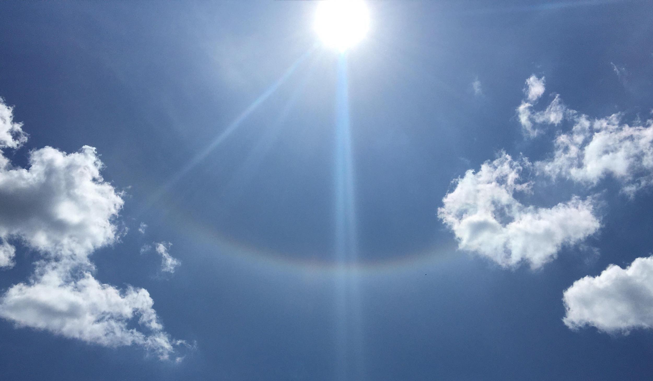 Sonne am blauen Himmel mit leichten Wolken und einem schwachen Lichtbogen