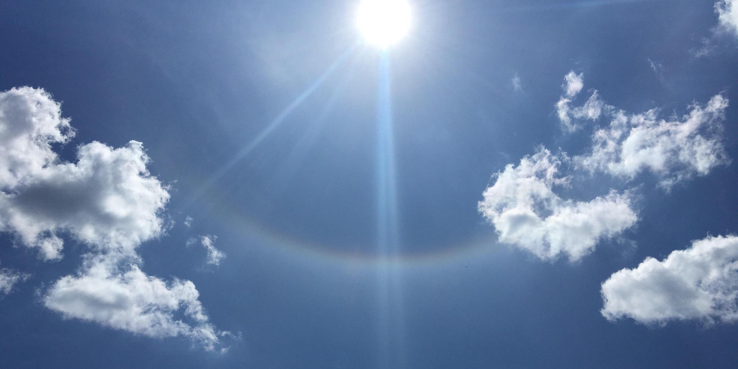 Sonne am blauen Himmel mit leichten Wolken und einem schwachen Lichtbogen