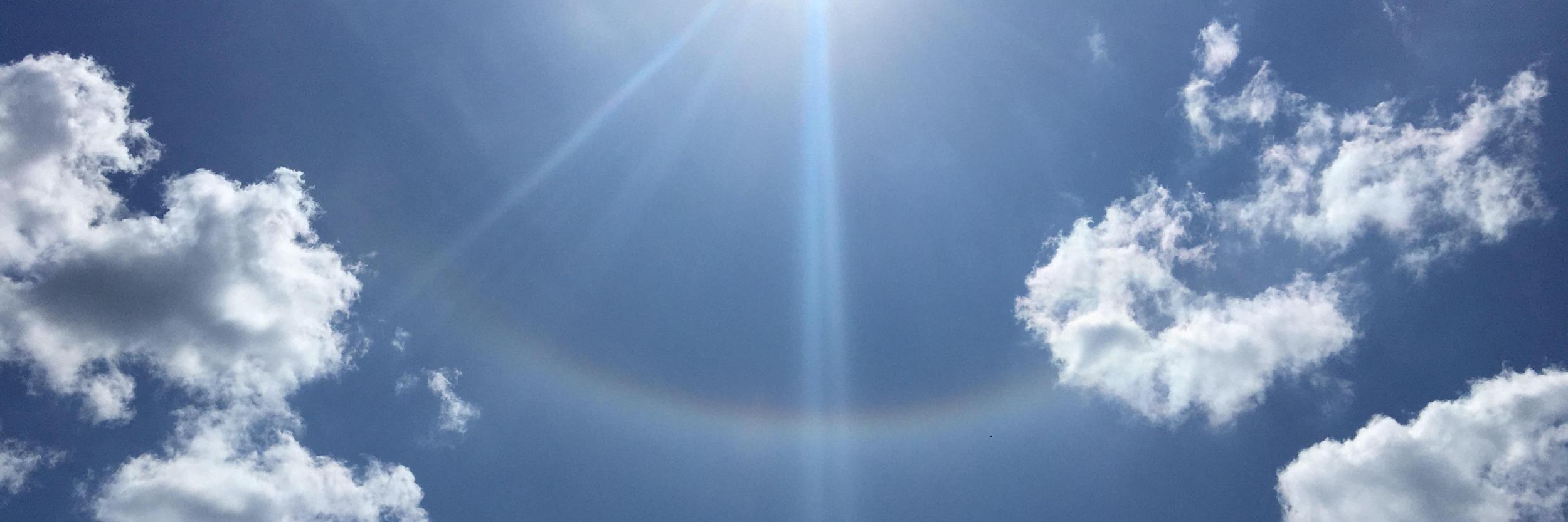 Sonne am blauen Himmel mit leichten Wolken und einem schwachen Lichtbogen