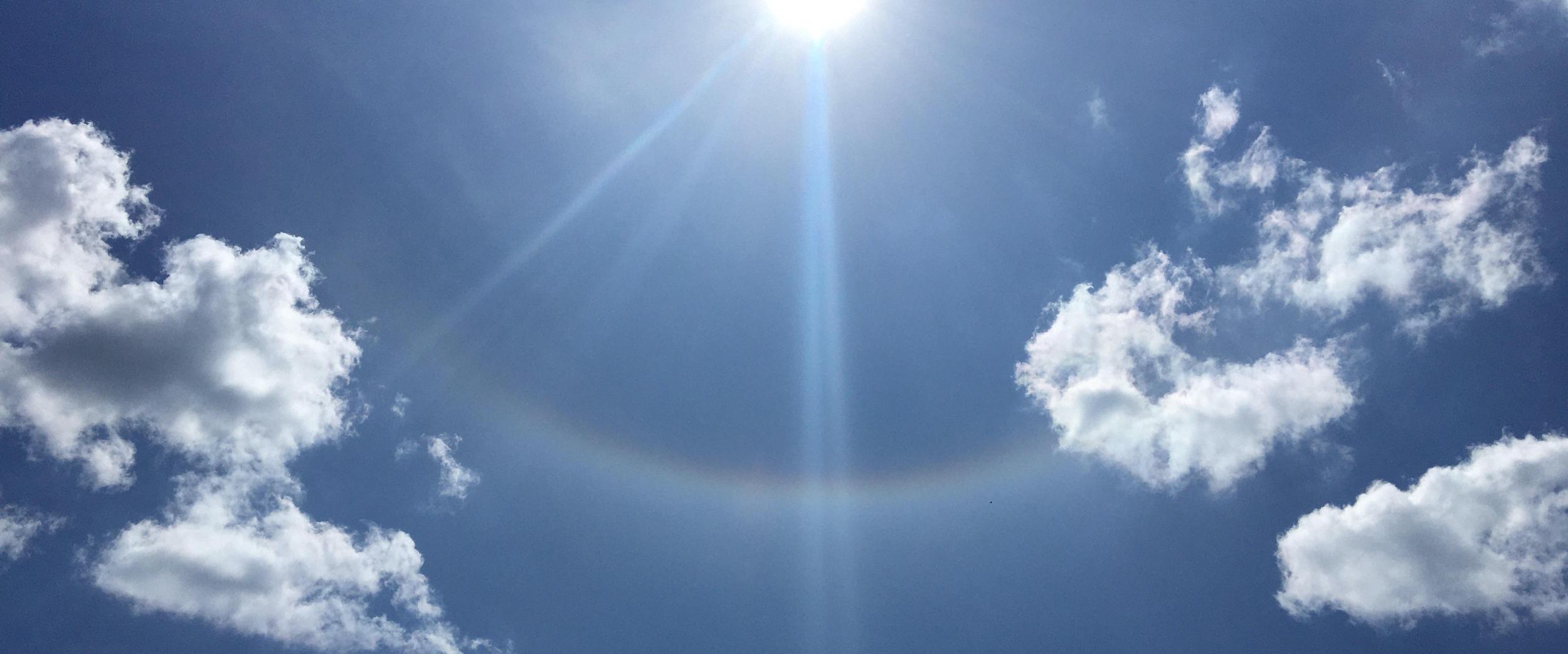 Sonne am blauen Himmel mit leichten Wolken und einem schwachen Lichtbogen