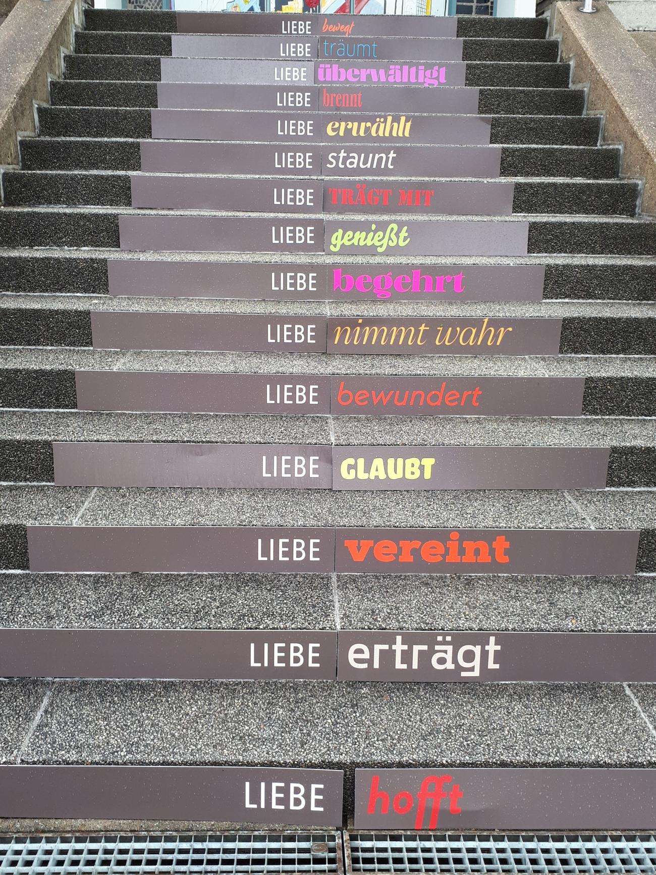 Steinerne Treppenstufen, auf deren Vorderseiten Schilder mit Textbotschaften angebracht sind. Jede Zeile beginnt mit dem Wort „LIEBE“, gefolgt von einem Verb in unterschiedlichen Farben und Schriftarten.