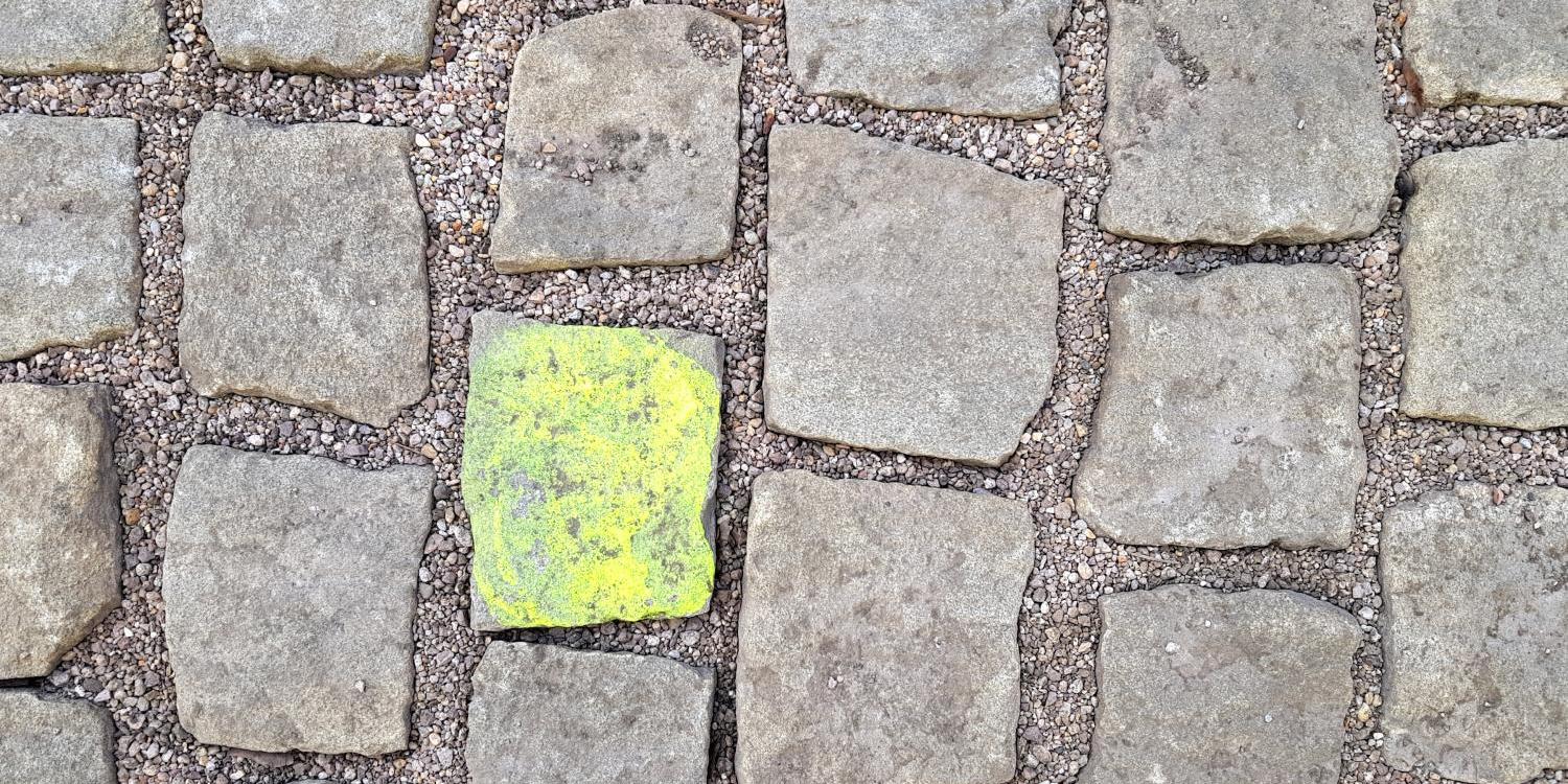Graues Kopfsteinpflaster mit einem einzelnen leuchtend gelben Stein in der Mitte.