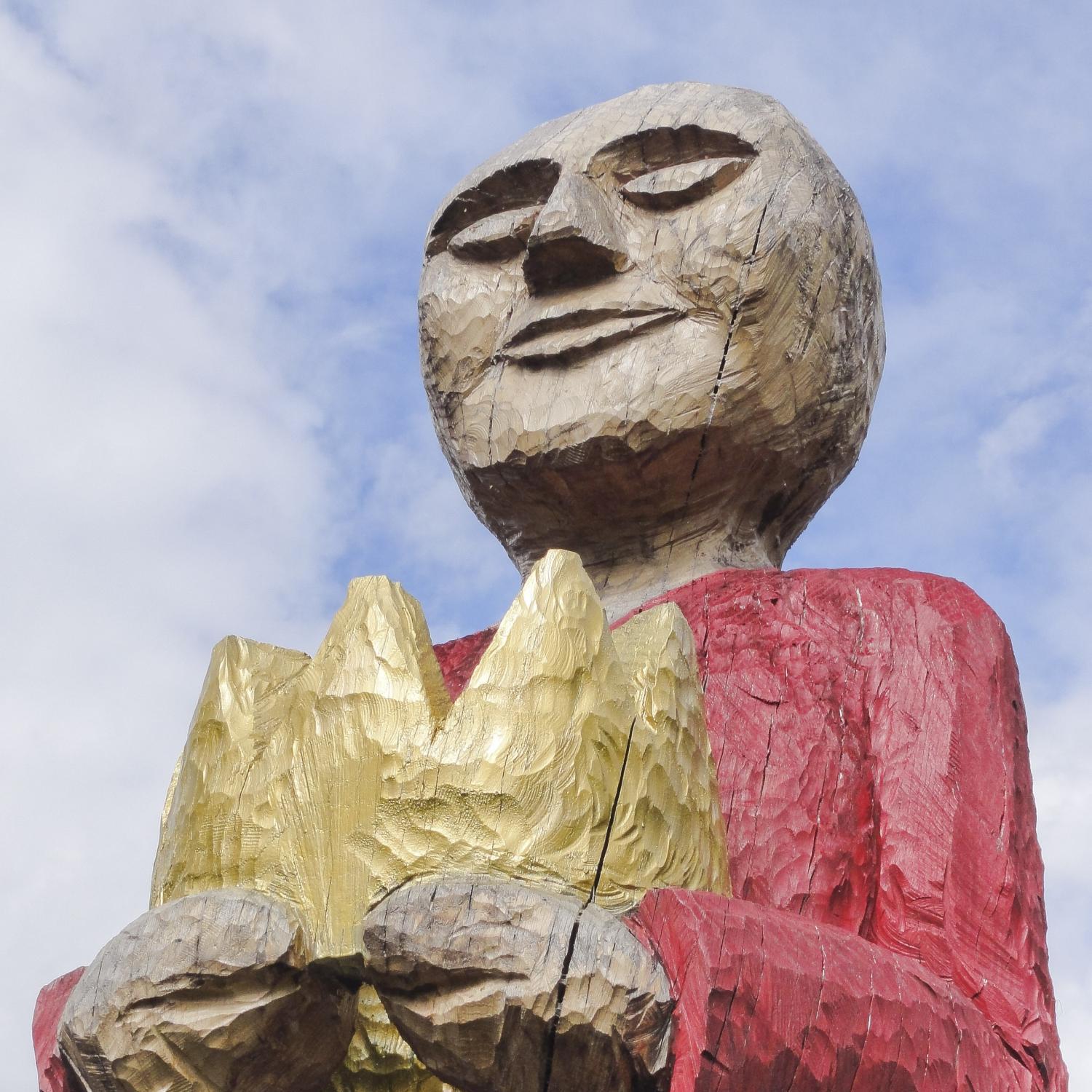 Holzskulptur einer Person mit rotem Oberteil, hält goldene Krone in den Händen.