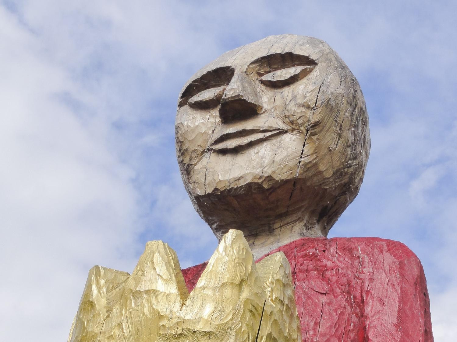 Holzskulptur einer Person mit rotem Oberteil, hält goldene Krone in den Händen.