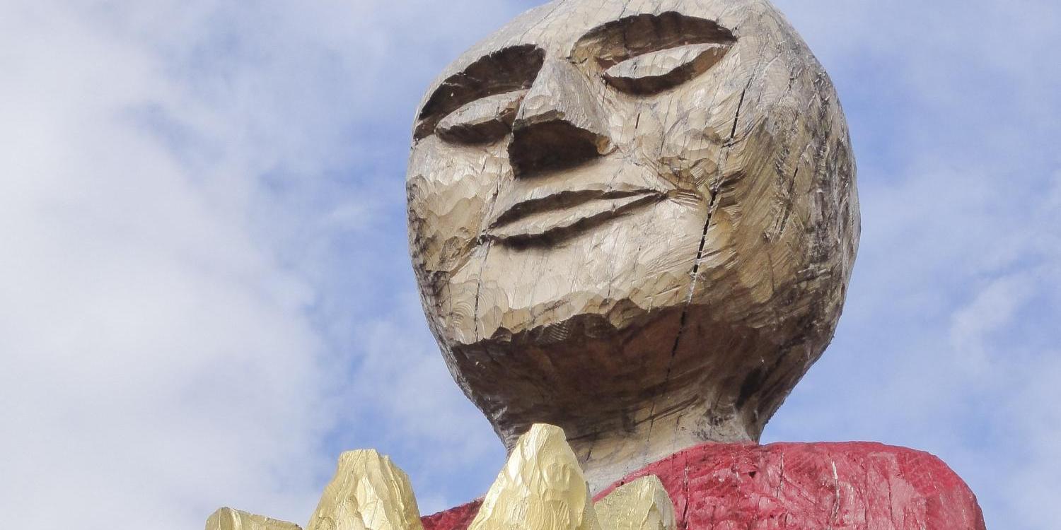 Holzskulptur einer Person mit rotem Oberteil, hält goldene Krone in den Händen.