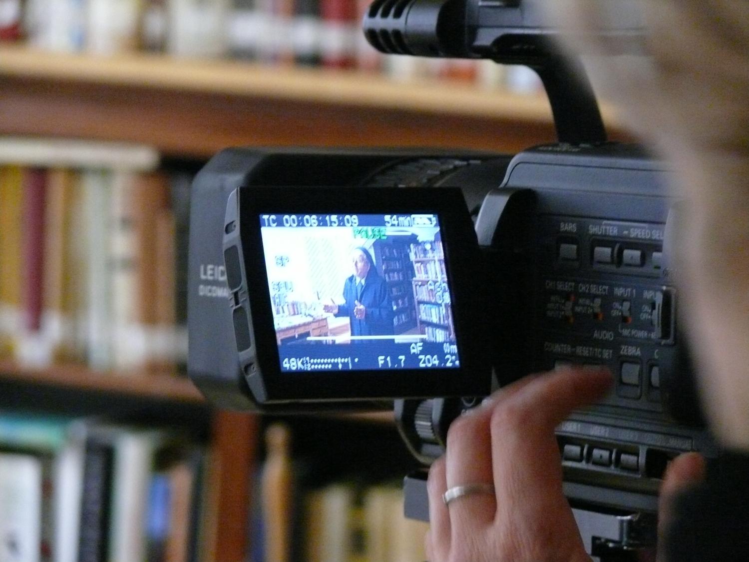 Nahaufnahme einer Videokamera, die eine Person vor Bücherregalen filmt.