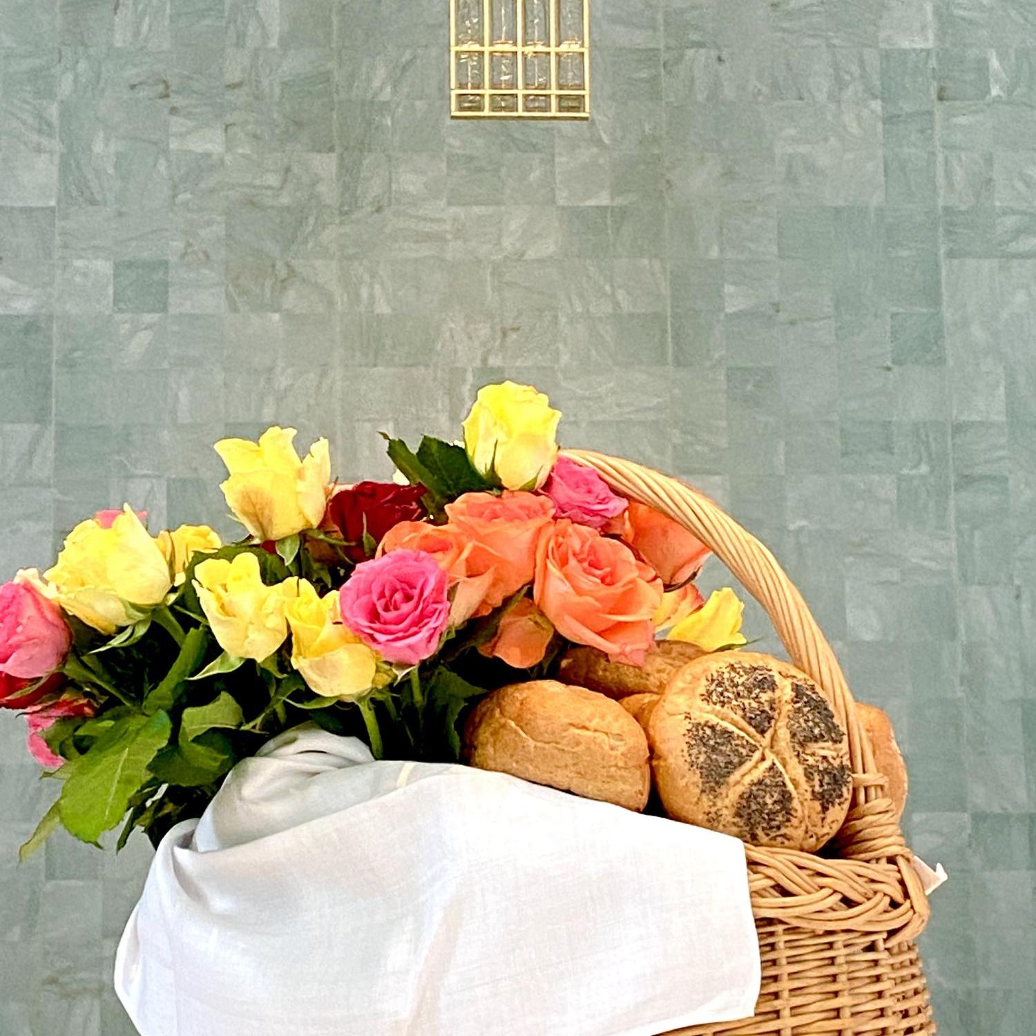 Korb mit Brötchen und bunten Rosen vor goldenem Kreuz an grauer Wand.