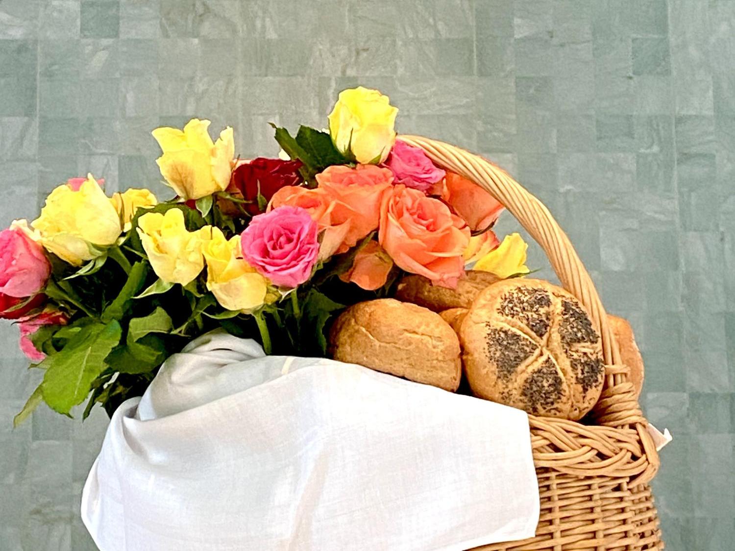 Korb mit Brötchen und bunten Rosen vor goldenem Kreuz an grauer Wand.