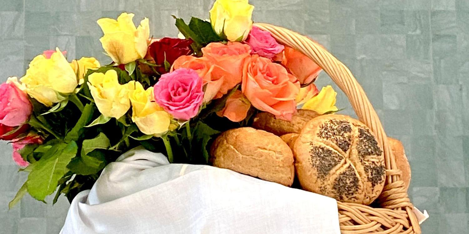 Rosen und Brot Korb mit Brötchen und bunten Rosen vor goldenem Kreuz an grauer Wand.