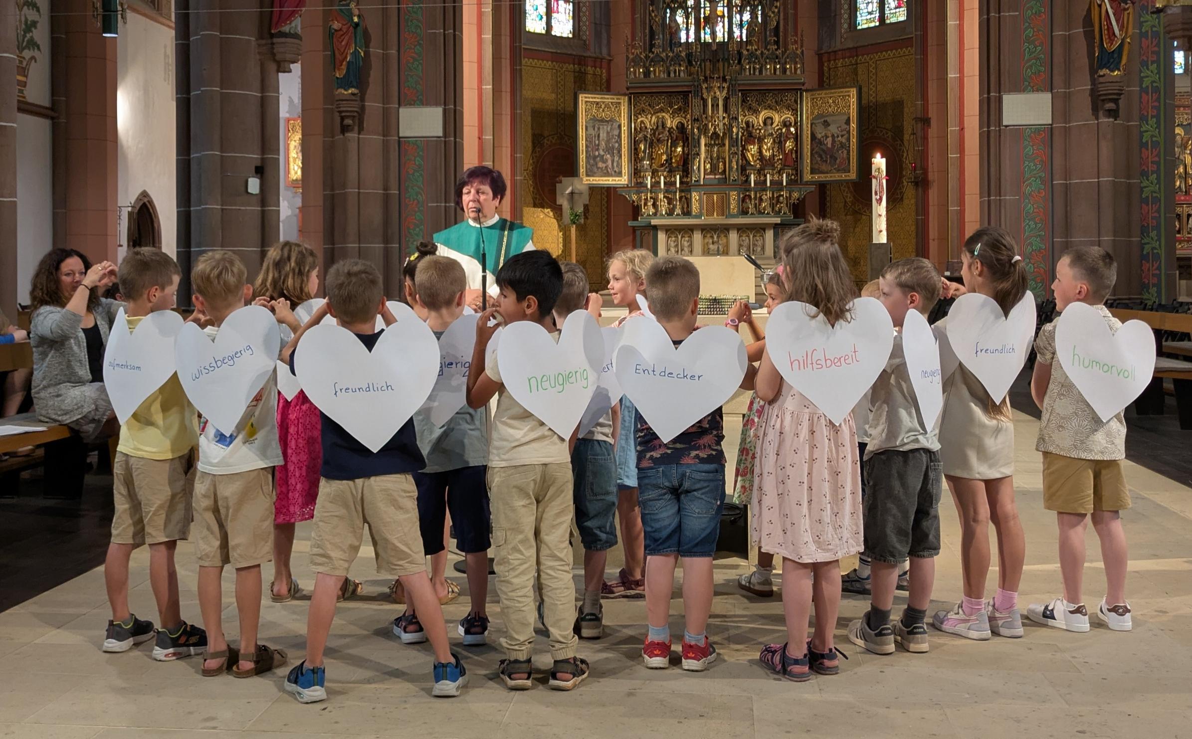 Kinder stehen mit dem Rücken zum Altar. Sie tragen Flügel in Herzform, auf dennen Eigenschaften stehen