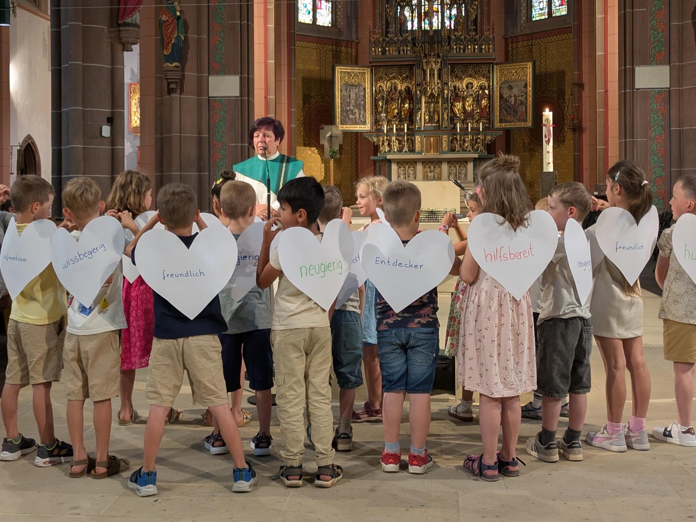 Kinder stehen mit dem Rücken zum Altar. Sie tragen Flügel in Herzform, auf dennen Eigenschaften stehen
