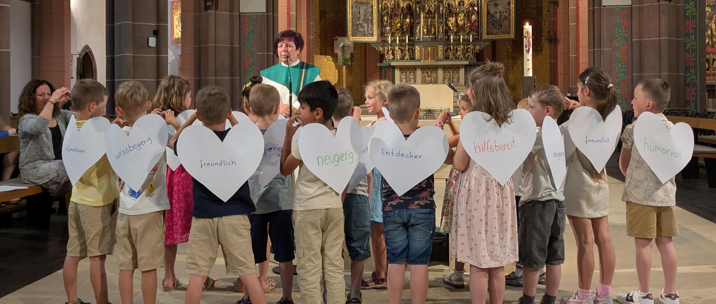 Kinder stehen mit dem Rücken zum Altar. Sie tragen Flügel in Herzform, auf dennen Eigenschaften stehen