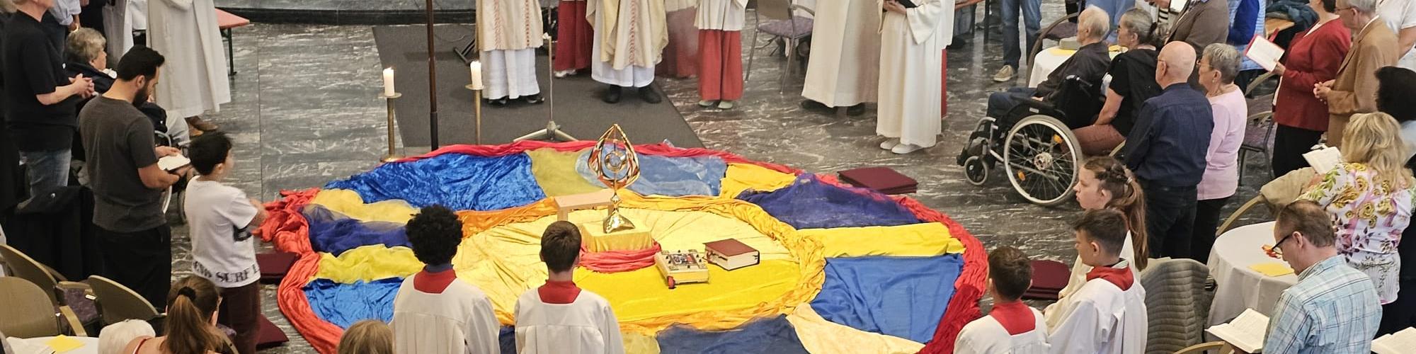 eine gelegte Mitte aus gelben, blauen und roten Tüchern. Darum versammelt Pfarrer Givens, Diakon Broessner, die Gemeindereferentinnen, Ministranten und Gläubigen