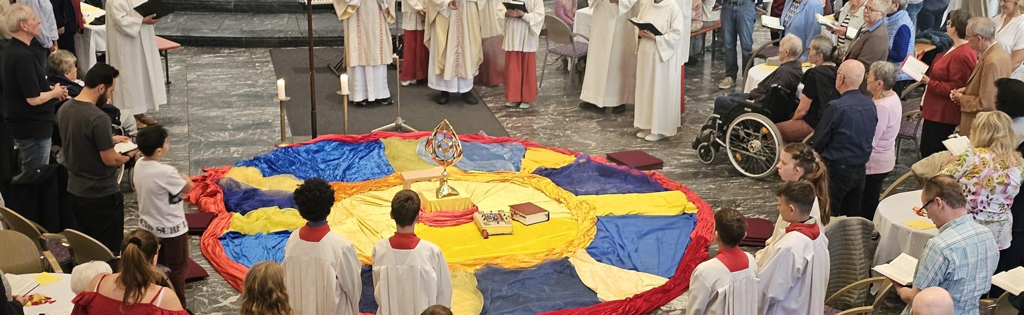 eine gelegte Mitte aus gelben, blauen und roten Tüchern. Darum versammelt Pfarrer Givens, Diakon Broessner, die Gemeindereferentinnen, Ministranten und Gläubigen