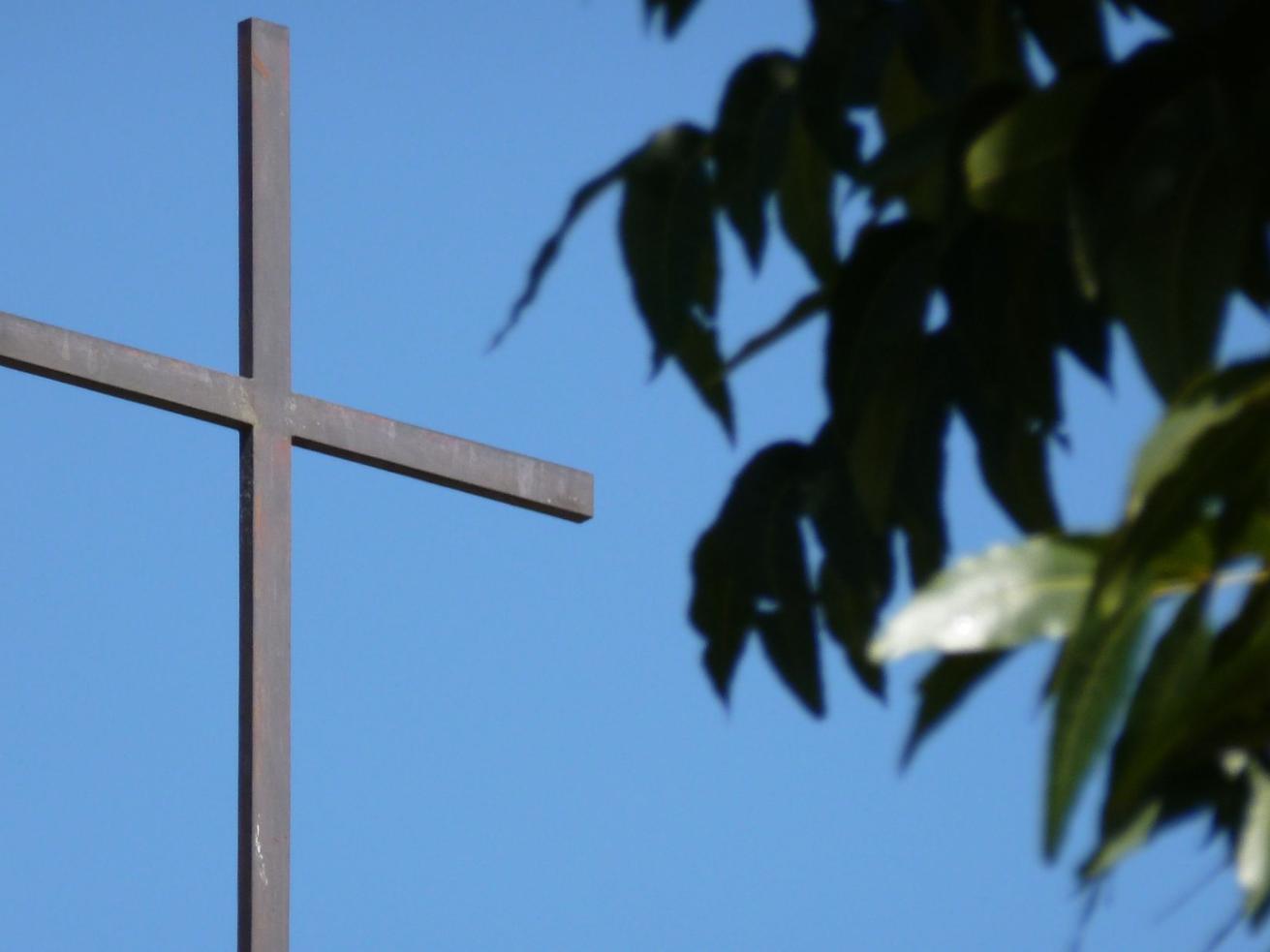 schlichtes Kreuz mit blauen Hinergrund. Rechts ragen Blätter an Ästen ins Bild