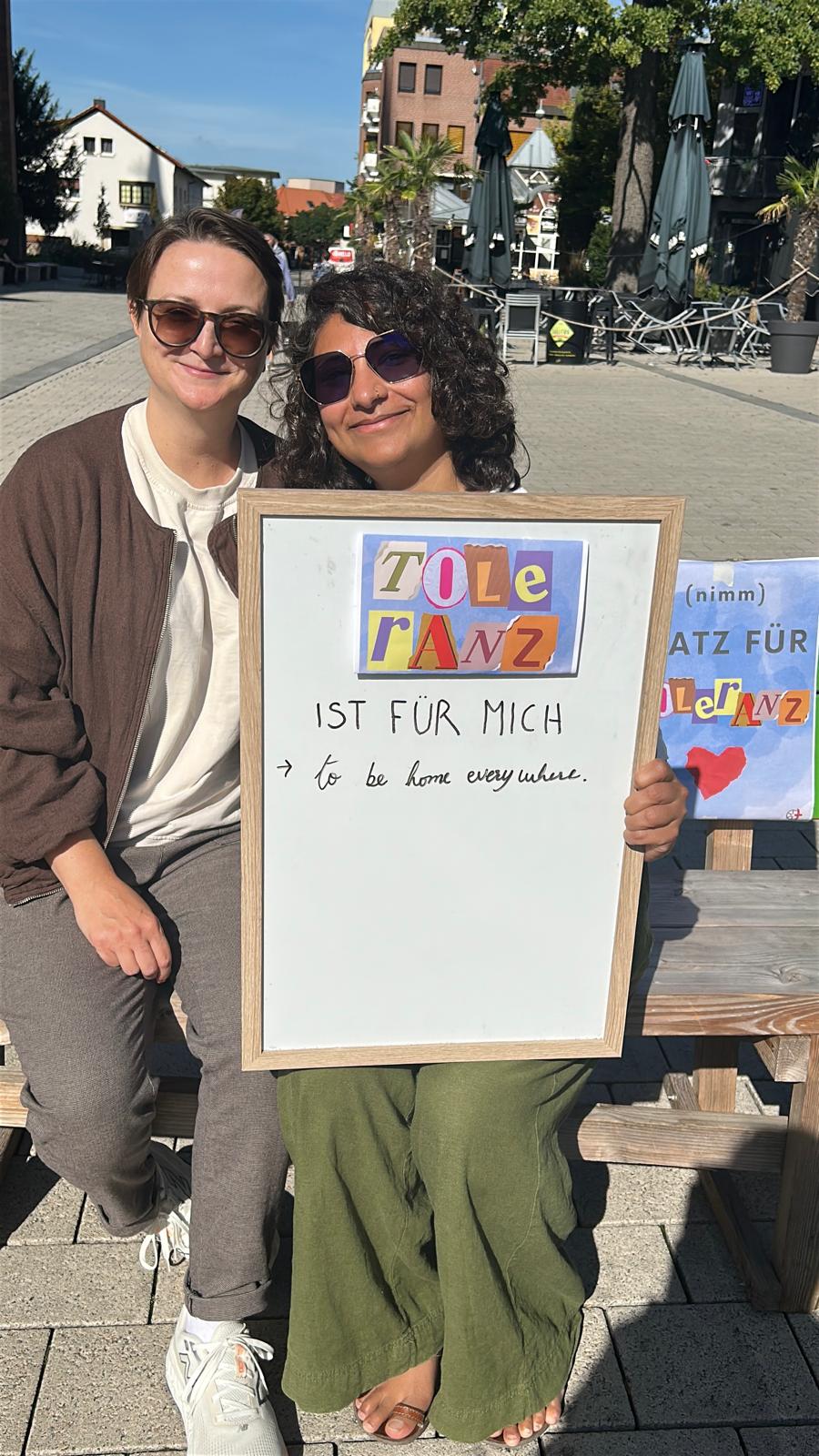 Zwei Frauen, sitzend auf der Babbelbank, halten ein Schild auf dem steht 'Toleranz ist für mich to be home everywhere'