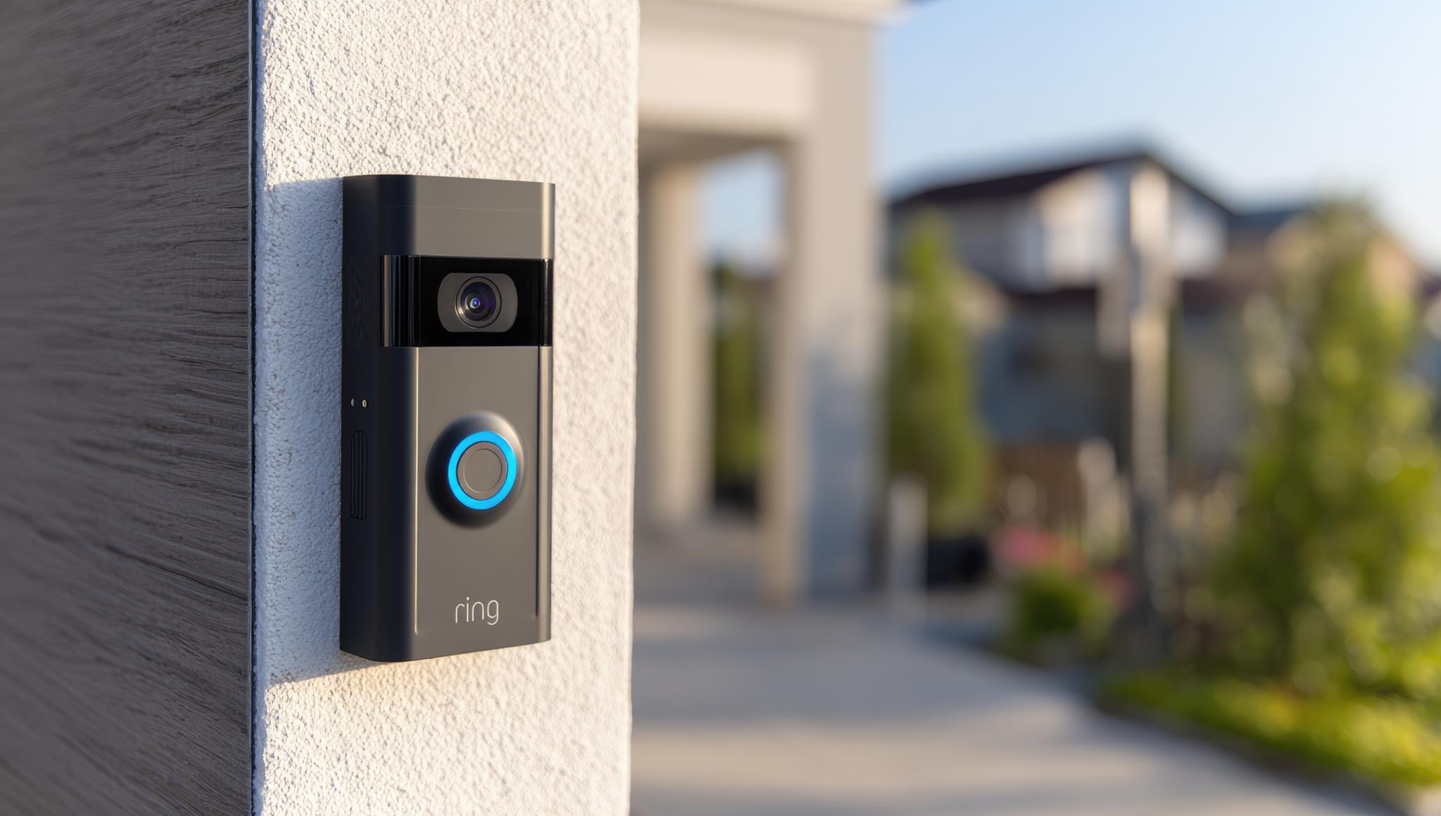 Modern doorbell camera mounted on light beige wall. Blurred background shows houses and trees