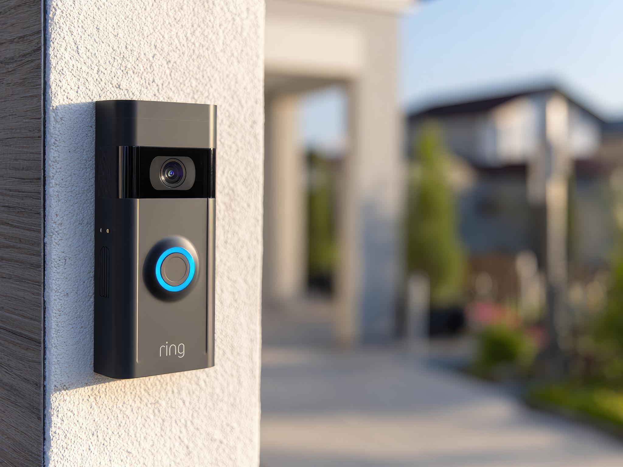 Modern doorbell camera mounted on light beige wall. Blurred background shows houses and trees