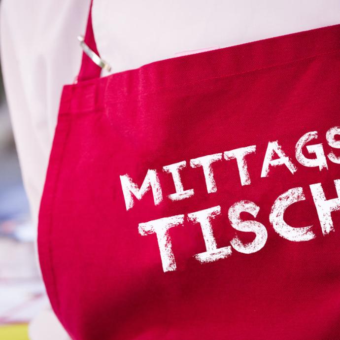Person mit roter Kochschürze mit der Aufschrift 'Mittagstisch'