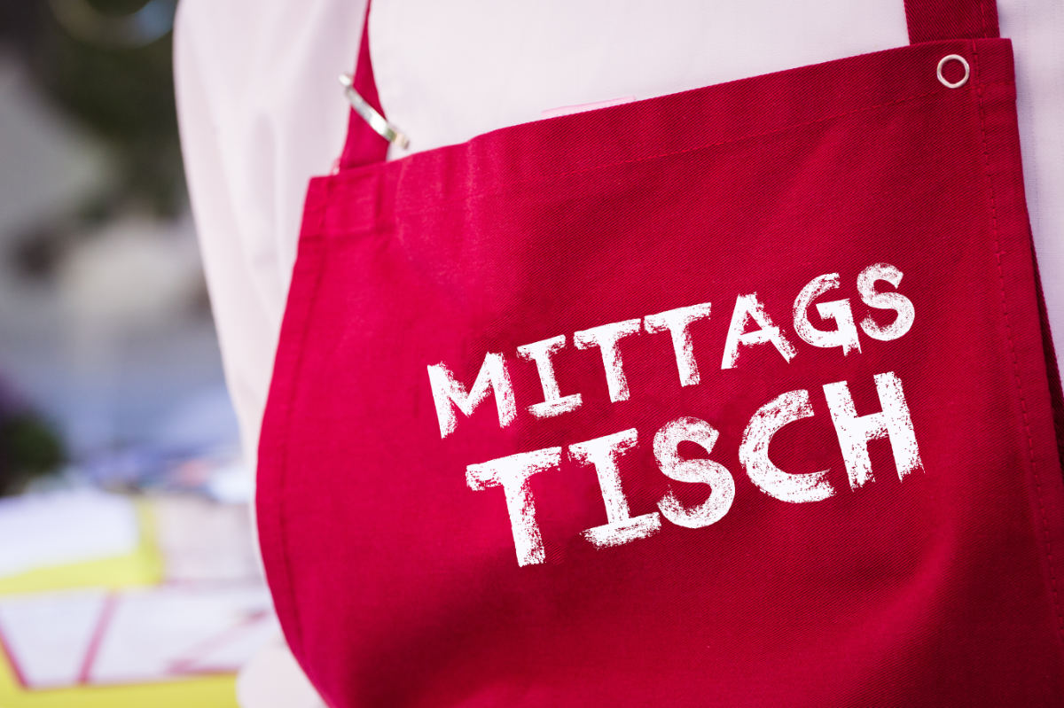 Person mit roter Kochschürze mit der Aufschrift 'Mittagstisch'